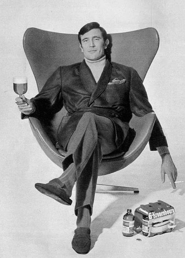 George Lazenby sentado en la silla del huevo de Arne Jacobsen con una Kronenbourg