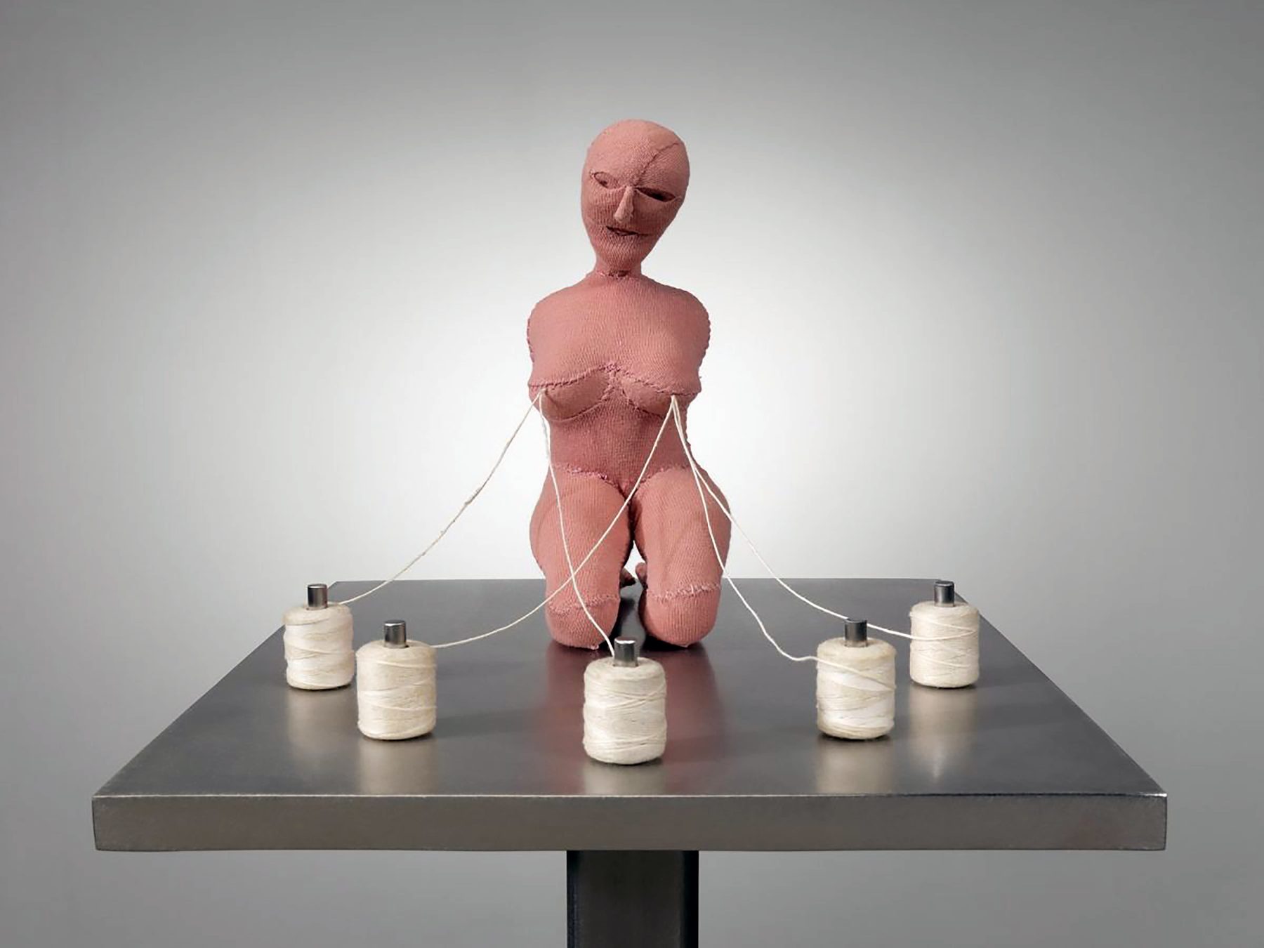 Pink fabric sculpture of a kneeling female figure on a metal table; white threads run from her breasts to five spools arranged around her.