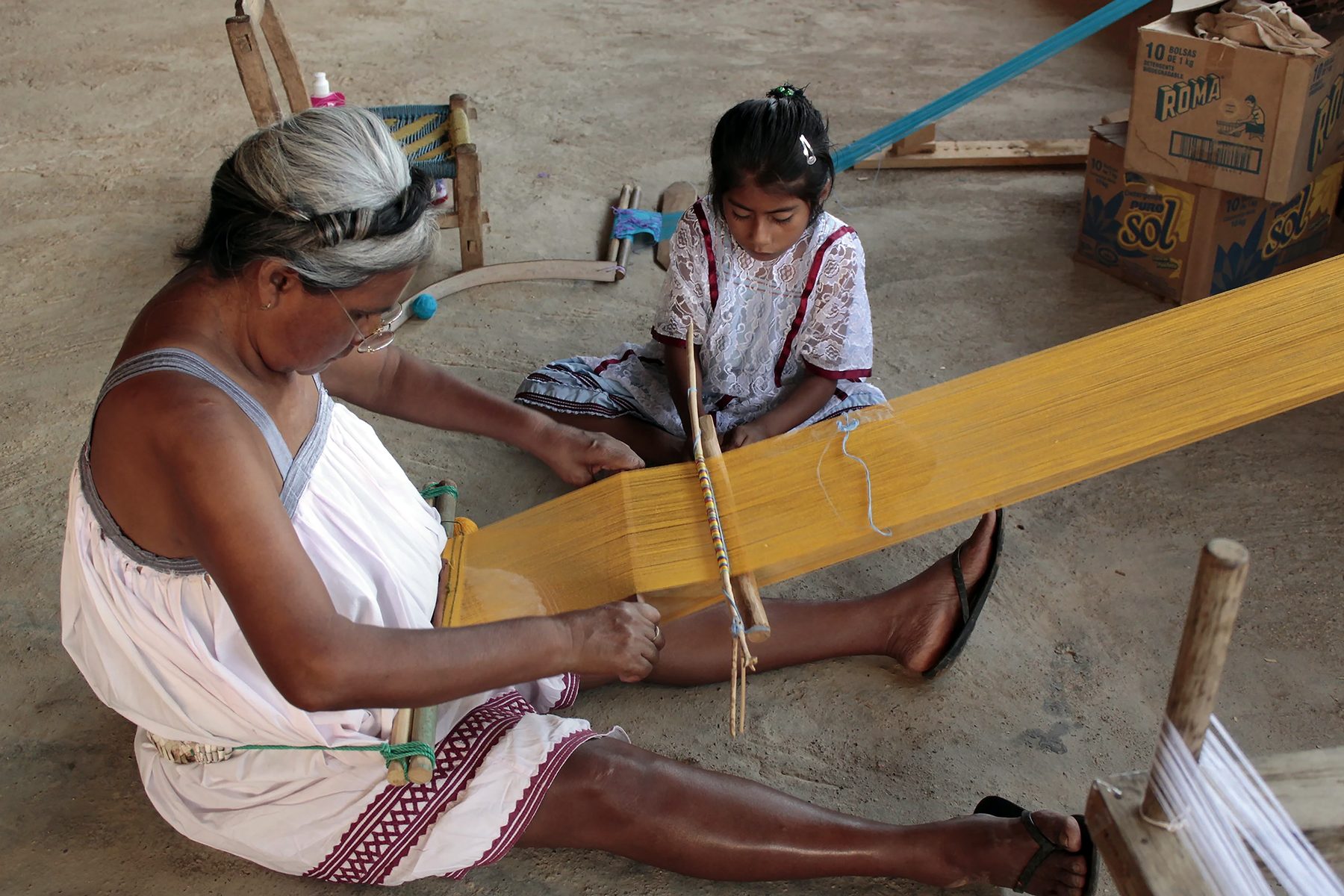 Indigenous woman sitting on the ground weaving on a backstrap loom with a yellow warp; beside her, a girl in a white dress watches.