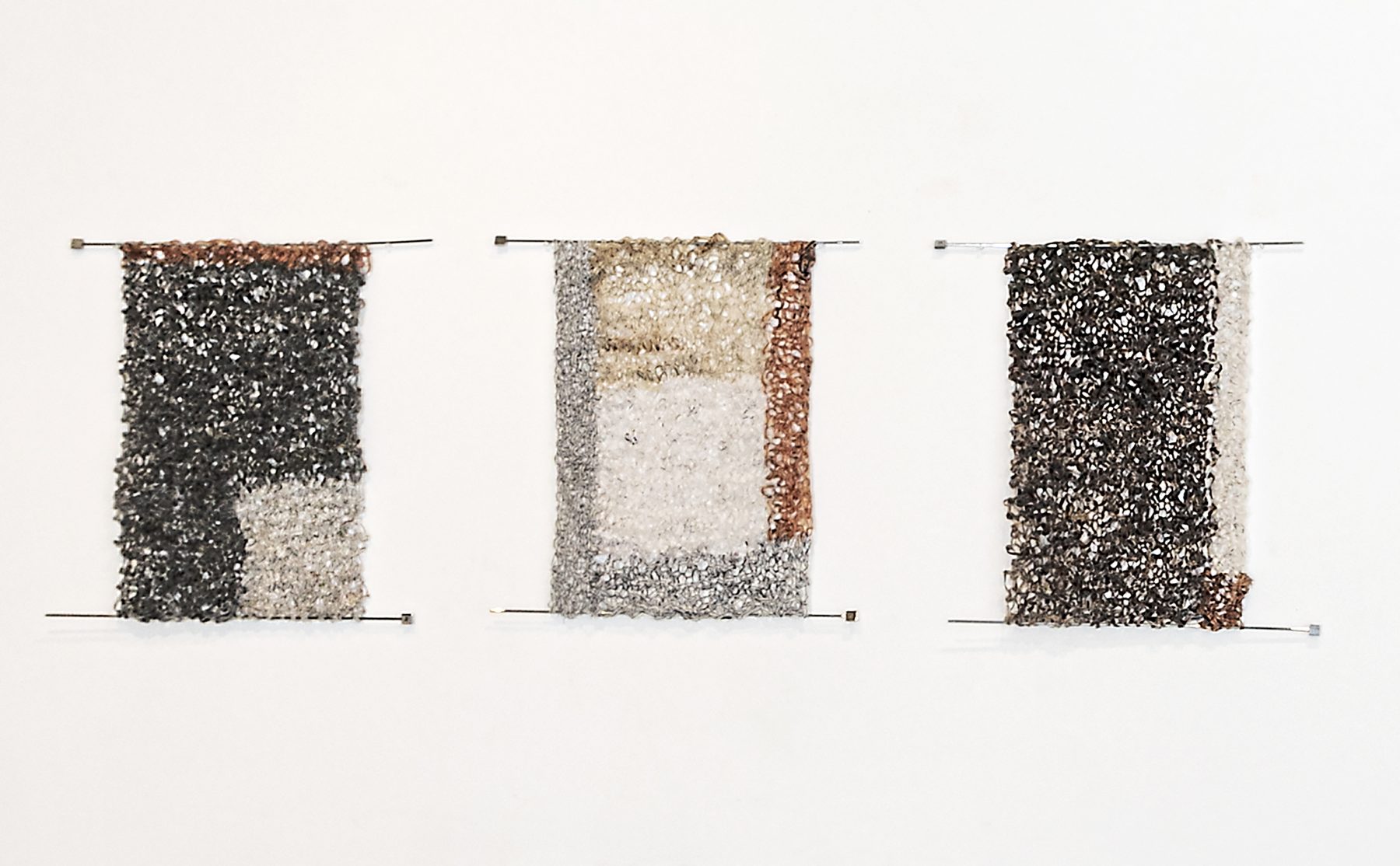 Three rectangular tapestries hanging on a white wall, woven from paper strips in black, grey, white and copper tones, each held by metal rods at the top and bottom.