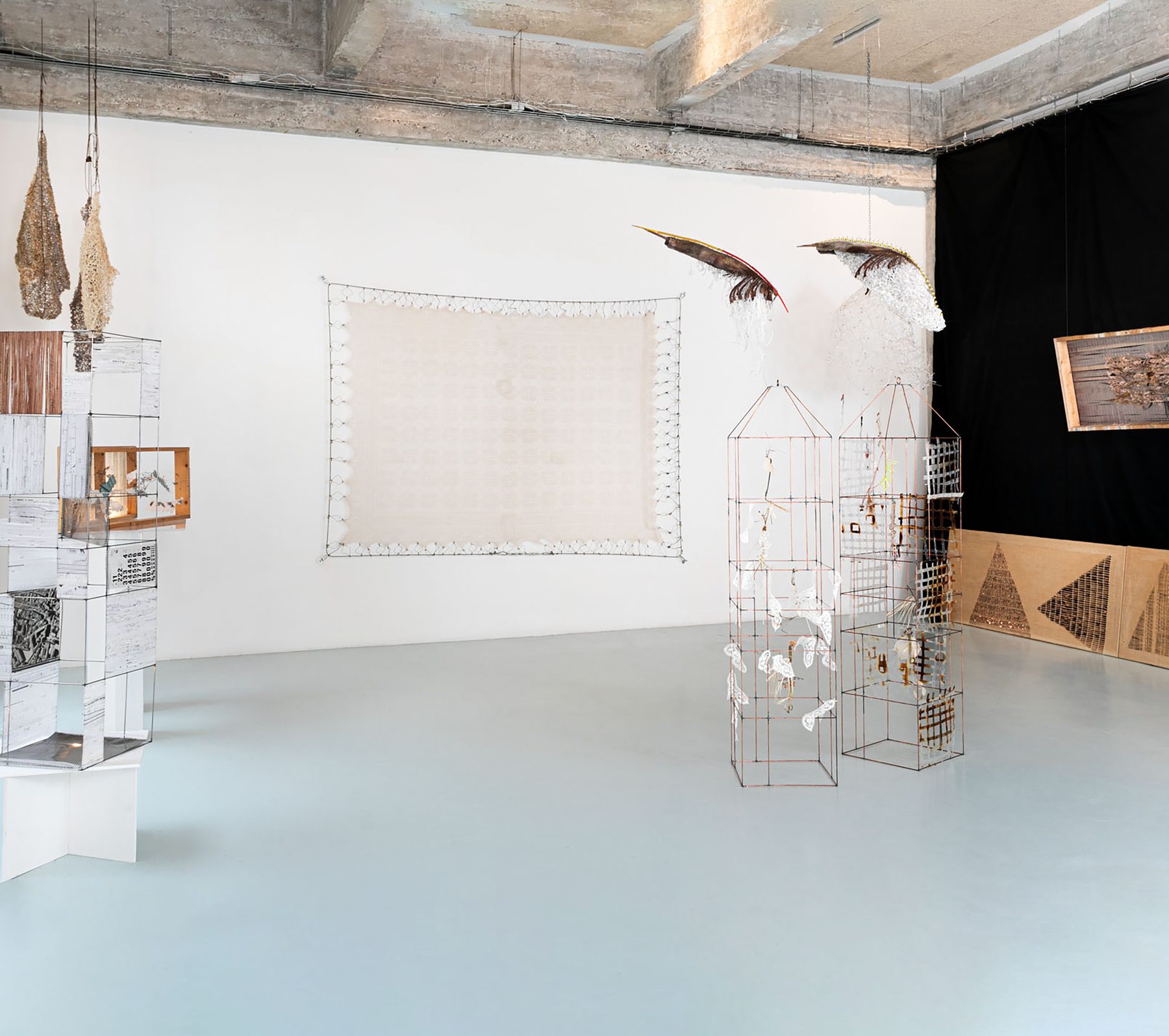 White exhibition room with a light grey floor: at the back, a large pale tapestry hangs on the wall; in front, wire cages holding paper pieces and, at the sides, hanging pieces and display cases with textiles.