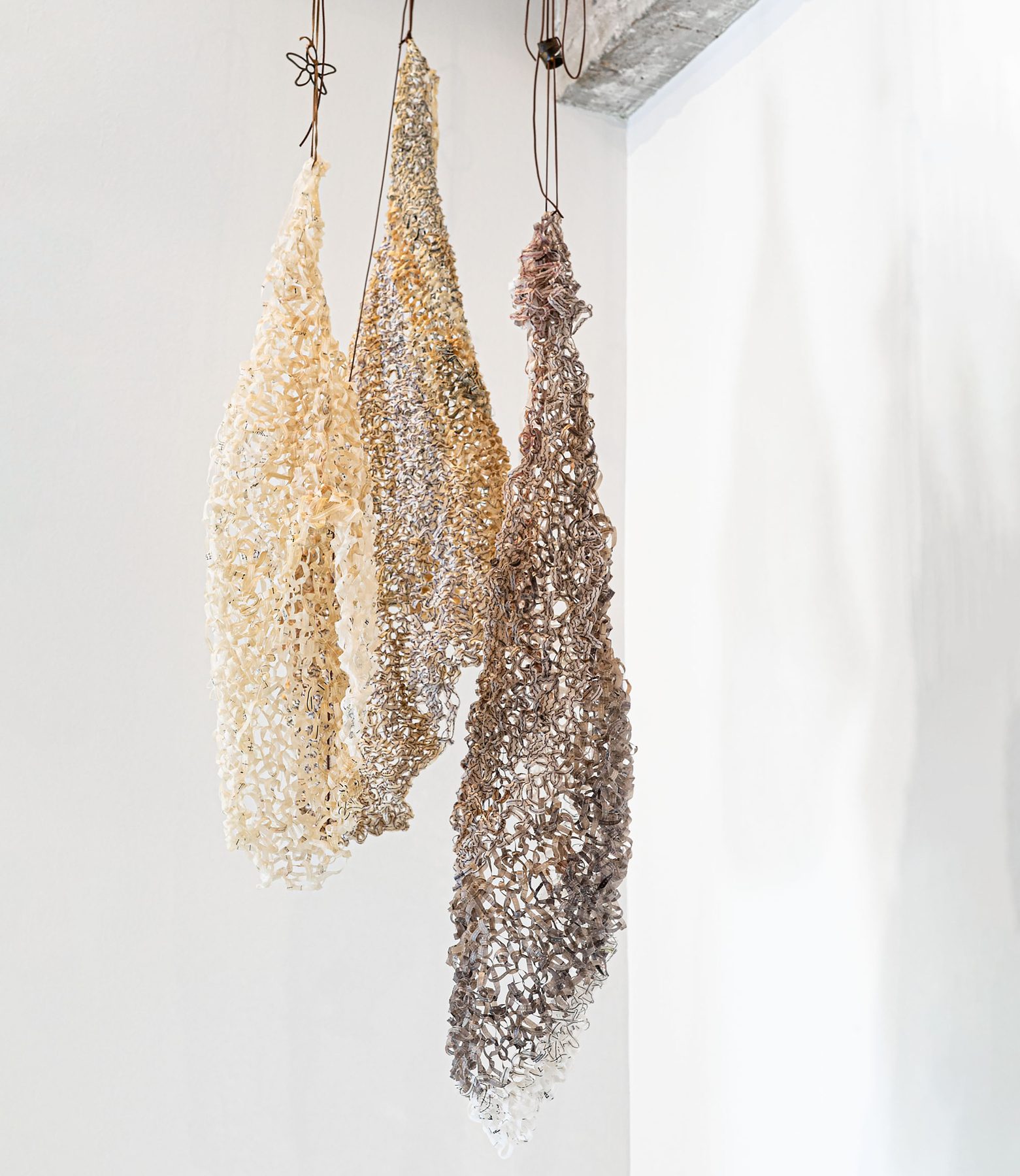 Three elongated, cocoon-shaped pieces knitted from paper thread in cream, gold and grey tones, hanging from the ceiling beside a white wall.
