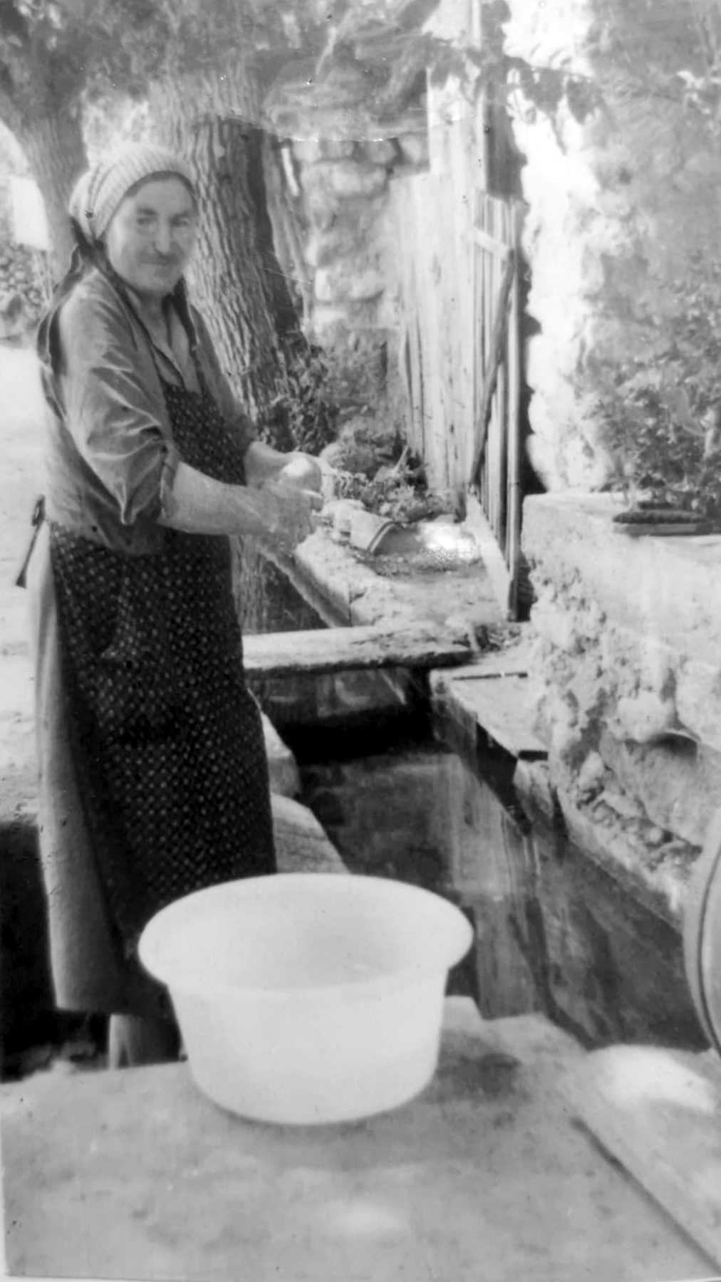Black-and-white photograph: an elderly woman wearing a headscarf and an apron washes clothes standing by the stone basin of a washhouse; in front of her, a white plastic tub.