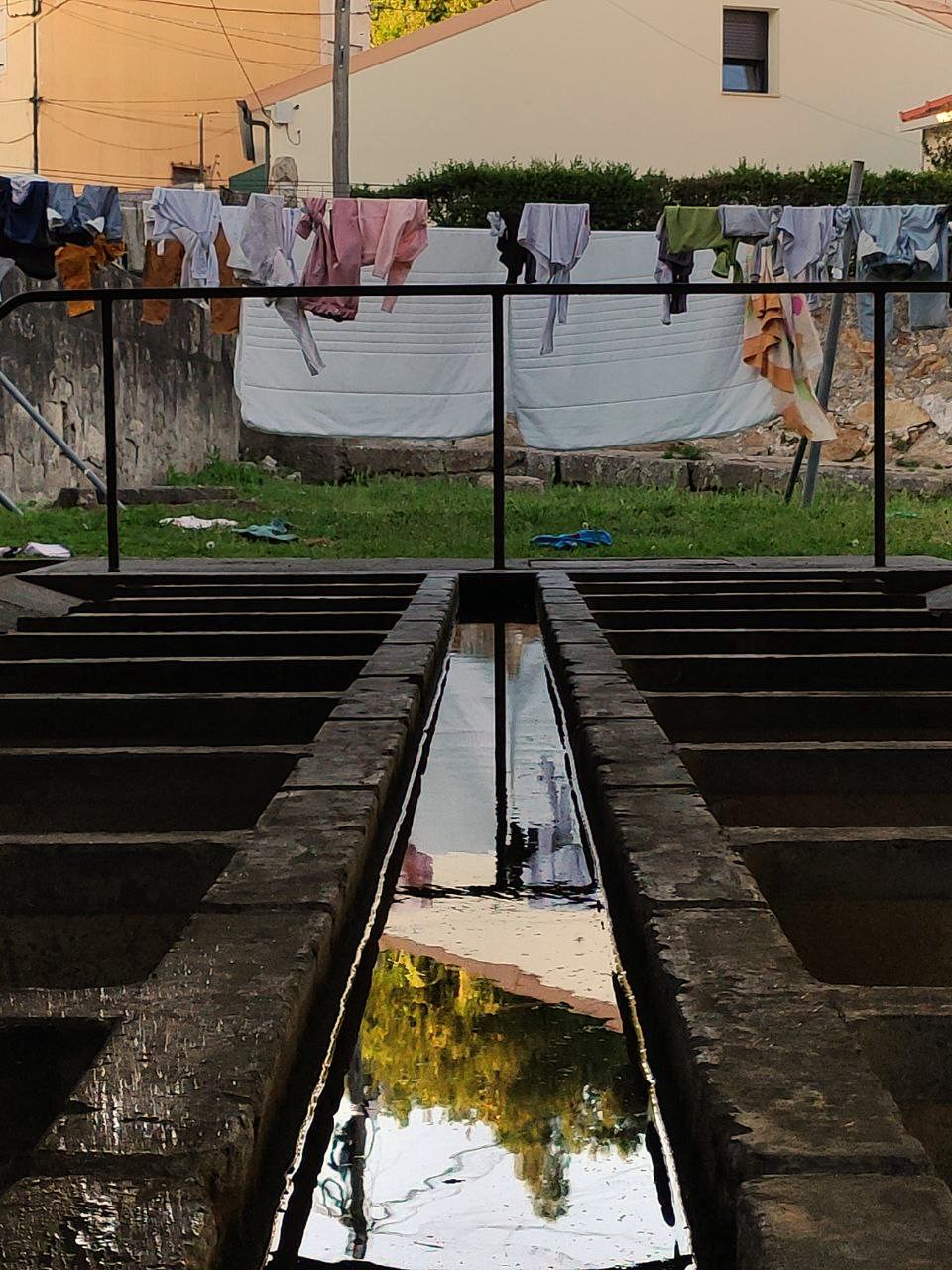Contumil washhouse: a central water channel between two rows of stone basins, reflecting the sky and trees; in the background, sheets and clothes hanging on lines above a railing.