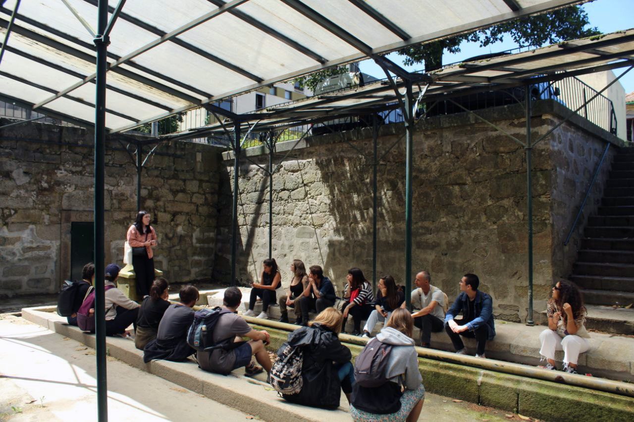 A group of people sitting on the edge of the basins of the Arca d’Água washhouse, under a translucent roof on green metal posts, listening to a standing woman, beside stone walls.