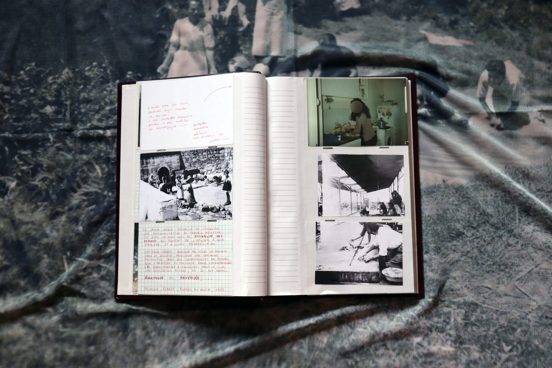An archive book lying open on a fabric printed with an old photograph: black-and-white archive photographs pasted onto its pages and handwritten notes in red.