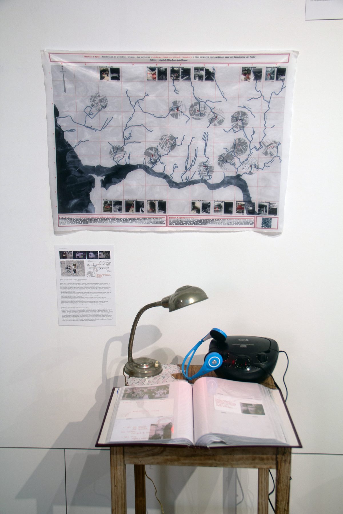 Installation in an exhibition room: on the wall, the cartography of the washhouses of Porto printed on fabric; below, a wooden table with the open archive book, a desk lamp, blue headphones and a CD player.