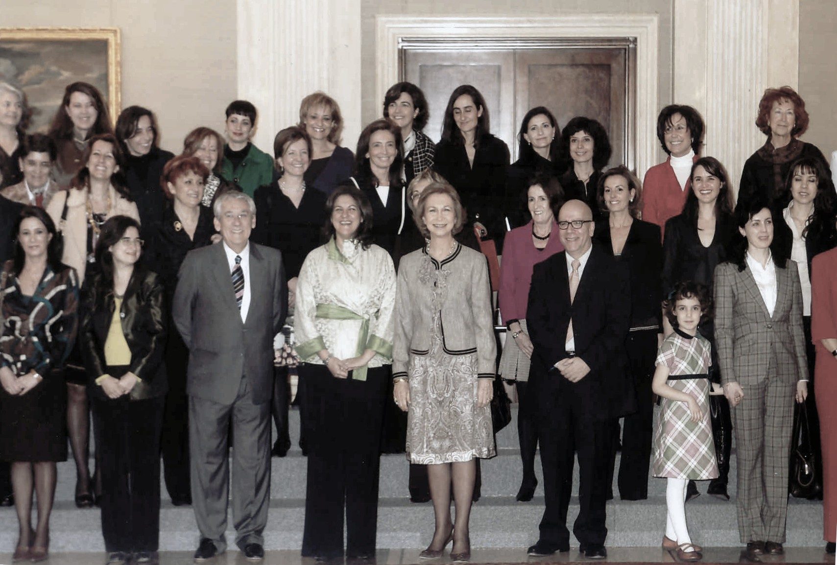 Fotografía de grupo en color: unas cuarenta mujeres, la mayoría vestidas de oscuro, posan en varias filas sobre una escalinata de un salón con columnas y una puerta de madera, junto a dos hombres y una niña en primera fila.