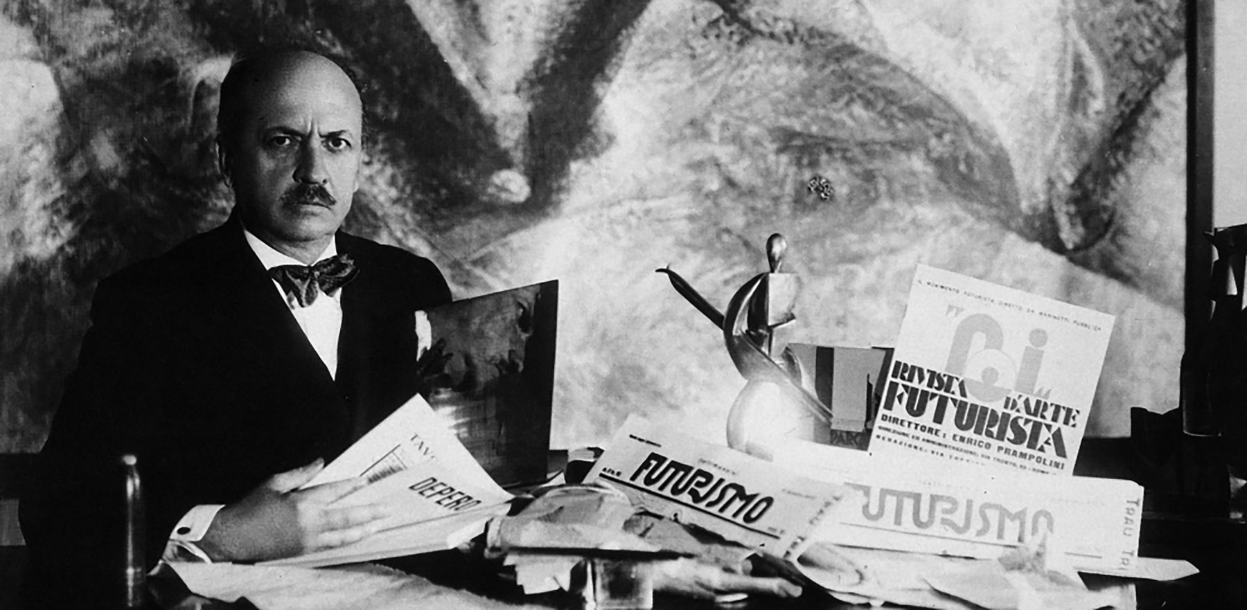 Fotografía en blanco y negro de Marinetti sentado a una mesa, con traje oscuro y pajarita, rodeado de periódicos y revistas futuristas y de una pequeña escultura metálica, ante un cuadro de fondo.